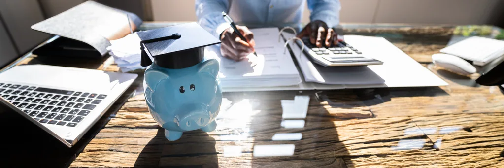 Piggy bank wearing a graduation cap beside a calculator and laptop, representing budgeting for the education assistant course costs in BC.