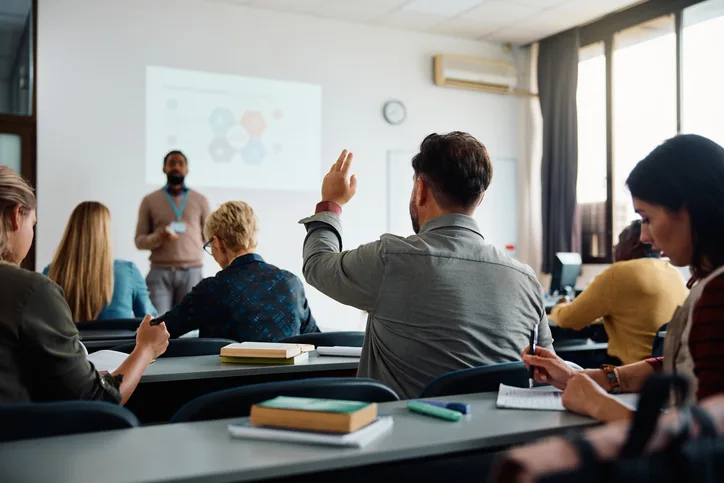Adult students in a classroom with one learner raising a hand during a lesson, representing education assistant training in BC.